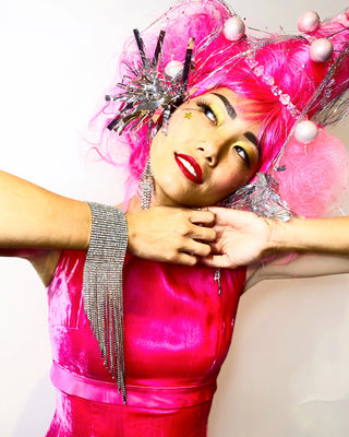 Woman wearing orange wig and color block dress holds up two fuchsia jewel earrings to her eyes