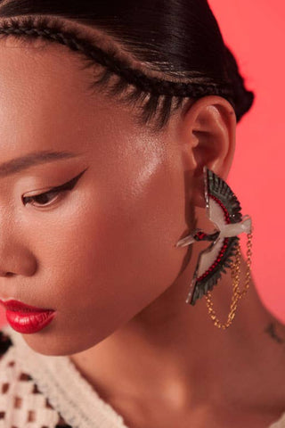 A person wearing a pair of silver crane earrings with black accents, set against a pink background.