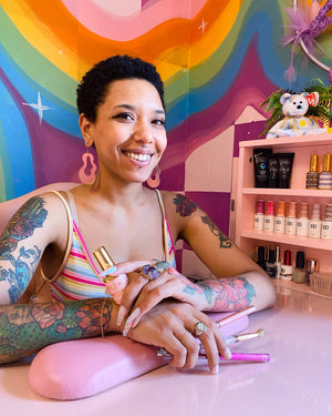 Nail tech smiles, sitting at a pink desk, in front of a colorful rainbow mural
