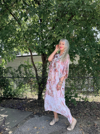 A woman with silver hair wearing a light pink and white floral kaftan dress walks outdoors on a gravel path under a leafy green tree.