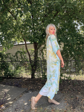 A woman with silver hair wearing a light blue and yellow tie-dye maxi dress, standing outdoors in a garden setting with trees and a fence in the background.