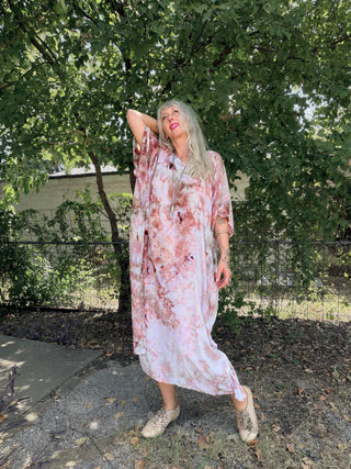 A woman wearing a loose, long-sleeve maxi dress with a pink and white floral pattern, standing outdoors in a park-like setting with trees and a fence behind her.