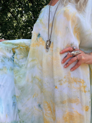 A close-up of a person wearing a flowing white kaftan-style dress with soft yellow and pale green tie-dye or watercolor washes. The person is outdoors with trees in the background, wearing a long silver pendant necklace and multiple rings.
