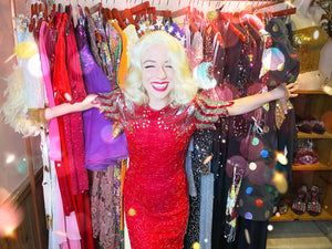 Sequin season! Woman wears red beaded sequin dress with star crown, smiling in front of a rack of party clothes