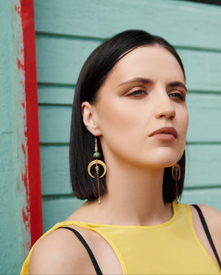 A woman with dark hair wearing gold crescent moon dangle earrings with green beads, posing in front of a light teal wooden wall with a red painted edge.