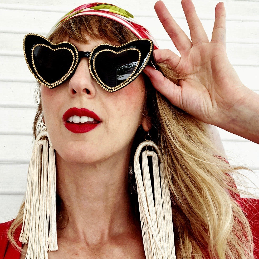 woman wears black cat eye heart shaped sunglasses trimmed in rhinestones and long tassel earrings