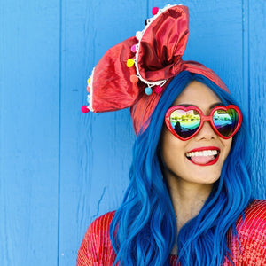 Woman wearing blue wavy wig and metallic red headwrap with colorful pompoms and red heart shaped frame sunglasses with rainbow mirror lens. She stands in front of a blue wall and smiles largely. She also wears a vintage red sequin blouse..